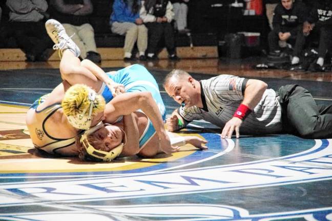 A wrestling official keeps a close watch as wrestlers grapple on the mat.